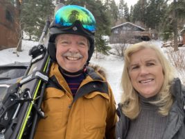 A man in skiing goggles and a yellow coat with a blonde woman in a grey sweater