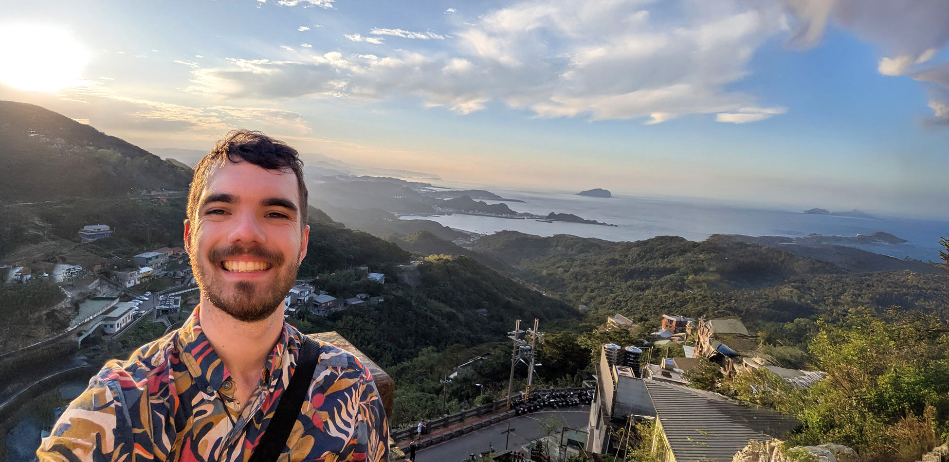 Patrick Hewes taking a selfie with a beautiful view of hills and sea in the background.